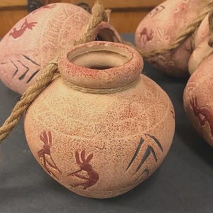 May include: A collection of small, terracotta-colored ceramic pots with a speckled finish. Each pot features a rope handle and is adorned with red and black tribal-style designs. The pots have a rustic, handmade appearance.