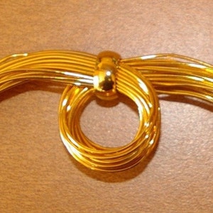 May include: A gold-colored brooch made of thin, interwoven wires forming a circle with two long, flowing strands of wire on either side.