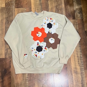 May include: A beige crewneck sweatshirt featuring three large floral-shaped appliqués made of fabric hexagons. The appliqués consist of solid orange, solid brown, and white floral-patterned fabric pieces. A small matching floral appliqué is on the sleeve.