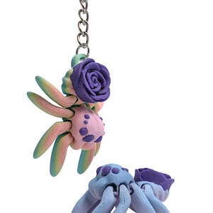 Puede incluir: Dos abalorios de araña de arcilla polimérica de colores con una flor morada en la parte superior. La araña de arriba es rosa, verde y azul con manchas moradas. La araña de abajo es azul con manchas moradas.