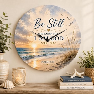 May include: A round wall clock featuring a beach sunset scene with tall grass and flying birds. The text reads Be Still AND KNOW THAT I AM GOD and PSALM 46:10 in dark blue lettering. The clock has black hands and a gold heart accent.