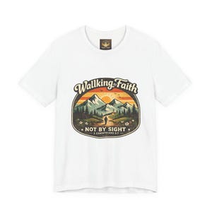 May include: White t-shirt with a graphic design. The design includes the text "Walking in Faith" above a mountain scene with a person walking on a path. The text "Not by Sight" and "2 Corinthians 5:7" are also included.