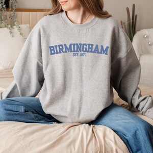 Könnte beinhalten: Ein grauer Pullover mit einem blauen Aufdruck, der "Birmingham Est. 1871" lautet.