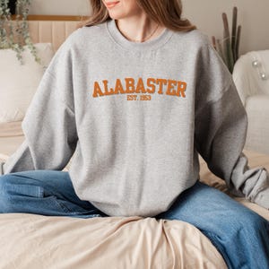 Könnte beinhalten: Ein grauer Pullover mit dem Text "ALABASTER EST. 1953" in Orange.