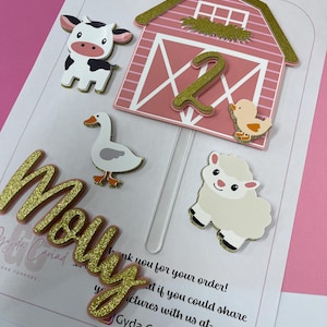May include: A farm-themed cake topper set with a pink barn, a cow, a sheep, a duck, a chick, and the number 2. The word "Molly" is included in gold glitter. The cake toppers are designed for a birthday celebration.