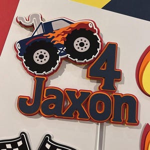 May include: A layered paper cake topper featuring a blue monster truck with orange flames, the number 4, and the name Jaxon in dark blue lettering with orange and red outlines. The design includes a white smoke trail and a white stick base.