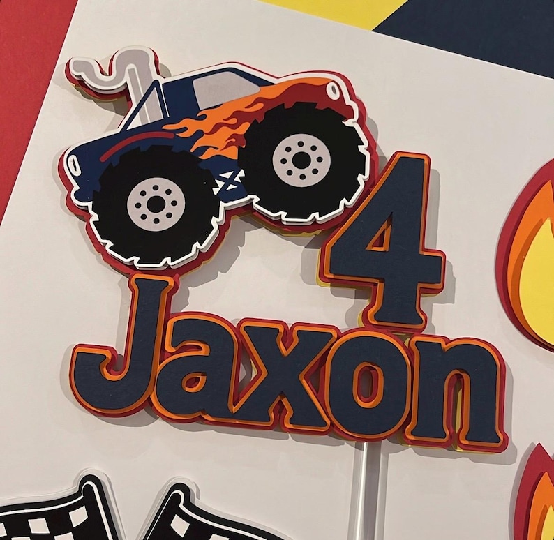 May include: A layered paper cake topper featuring a blue monster truck with orange flames, the number 4, and the name Jaxon in dark blue lettering with orange and red outlines. The design includes a white smoke trail and a white stick base.
