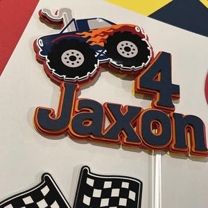 May include: A layered paper cake topper featuring a dark blue monster truck with orange flames, the name Jaxon in dark blue letters, and the number 4 in dark blue, all with orange and yellow accents, accompanied by two black and white checkered flags.