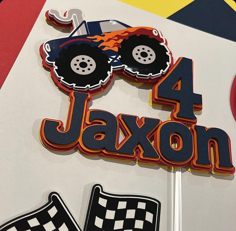 May include: A layered paper cake topper featuring a dark blue monster truck with orange flames, the name Jaxon in dark blue letters, and the number 4 in dark blue, all with orange and yellow accents, accompanied by two black and white checkered flags.