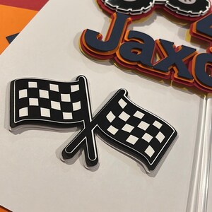 May include: A layered paper cake topper featuring the name Jaxon in dark blue, orange, and yellow, topped with a monster truck graphic. Below it, a separate cutout shows two crossed black and white checkered racing flags on a clear stick.
