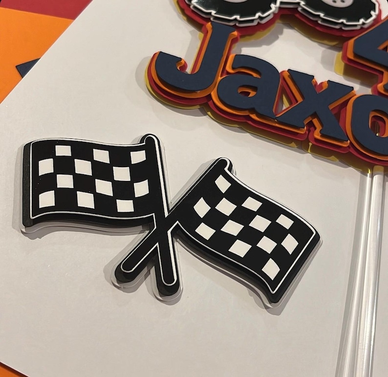 May include: A layered paper cake topper featuring the name Jaxon in dark blue, orange, and yellow, topped with a monster truck graphic. Below it, a separate cutout shows two crossed black and white checkered racing flags on a clear stick.