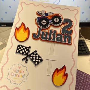 May include: A layered cake topper set featuring a blue monster truck with orange flames, the name Julian in dark gray, the number 2, two checkered racing flags, and two yellow and orange flame cutouts, all attached to a clear plastic stick.