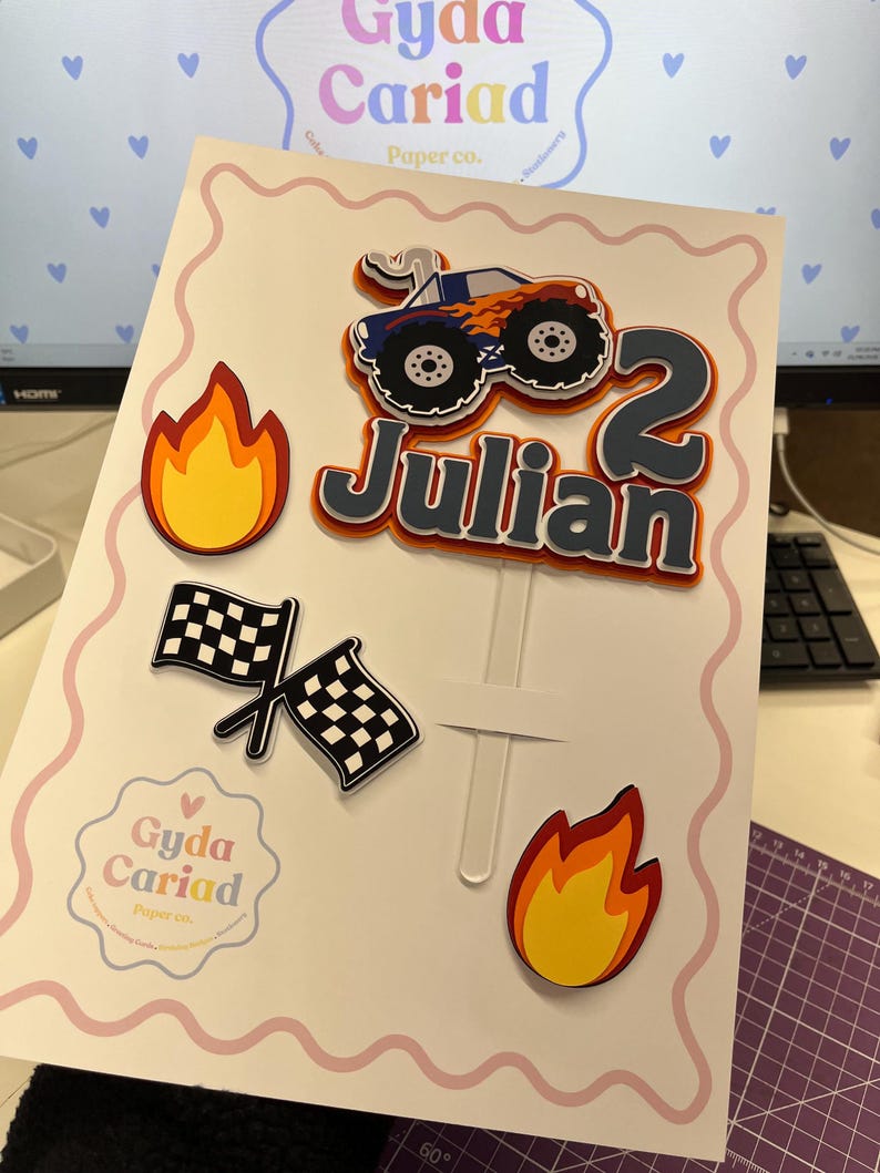 May include: A layered cake topper set featuring a blue monster truck with orange flames, the name Julian in dark gray, the number 2, two checkered racing flags, and two yellow and orange flame cutouts, all attached to a clear plastic stick.