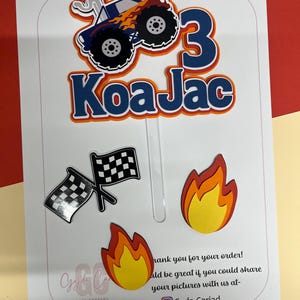 May include: A cake topper set featuring a blue monster truck with orange flames, the number 3, and the name Koa Jac in blue and orange. Includes two separate flame cutouts and a pair of black and white checkered racing flags on clear sticks.