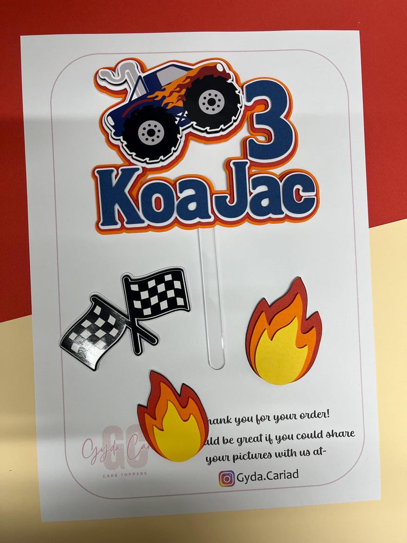 May include: A cake topper set featuring a blue monster truck with orange flames, the number 3, and the name Koa Jac in blue and orange. Includes two separate flame cutouts and a pair of black and white checkered racing flags on clear sticks.