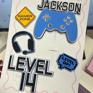 May include: A gaming-themed cake topper set featuring a blue game controller, a black headset, a yellow diamond-shaped sign reading GAMER ZONE, and text reading JACKSON, LEVEL 14, and LEVEL UP! in a pixelated blue speech bubble design.