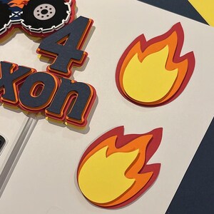 May include: A layered paper cake topper featuring a monster truck graphic, the number 4, and the name Nixon in dark blue, orange, and yellow. Two separate flame cutouts in yellow, orange, and red are included as decorative accents for the display.