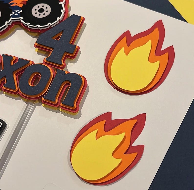 May include: A layered paper cake topper featuring a monster truck graphic, the number 4, and the name Nixon in dark blue, orange, and yellow. Two separate flame cutouts in yellow, orange, and red are included as decorative accents for the display.