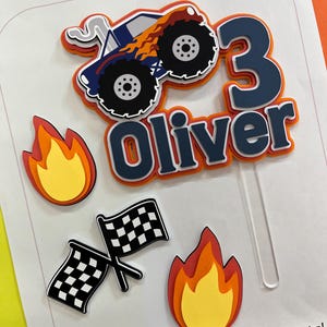 May include: A layered paper cake topper featuring a blue monster truck with orange flames, the number 3, and the name Oliver in dark blue letters. The set includes two separate flame cutouts and two crossed black and white checkered racing flags.