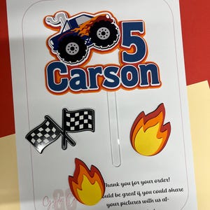 May include: A set of paper cake toppers featuring a blue monster truck with orange flames, the number 5, the name Carson in blue, two crossed black and white checkered flags, and two yellow and orange flame cutouts on a clear plastic stick.