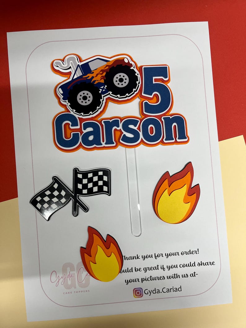 May include: A set of paper cake toppers featuring a blue monster truck with orange flames, the number 5, the name Carson in blue, two crossed black and white checkered flags, and two yellow and orange flame cutouts on a clear plastic stick.
