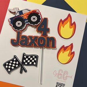 May include: A layered cake topper set featuring a blue monster truck with orange flames, the number 4, the name Jaxon in dark blue, two yellow and orange flame cutouts, and two black and white checkered flags on a clear plastic stick.