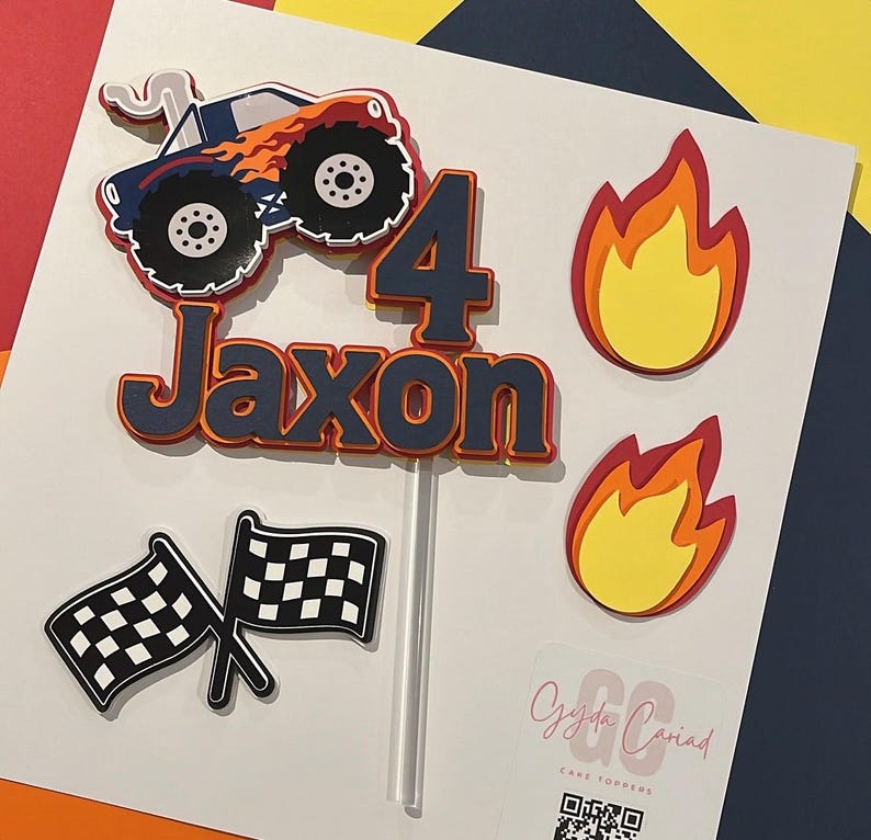 May include: A layered cake topper set featuring a blue monster truck with orange flames, the number 4, the name Jaxon in dark blue, two yellow and orange flame cutouts, and two black and white checkered flags on a clear plastic stick.