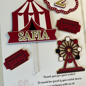 May include: A set of circus-themed cake toppers including a red and white striped tent with the name SAFIA, a scalloped sign with the number 2, two red circus admit one tickets, and a ferris wheel, all featuring red, white, and gold accents.