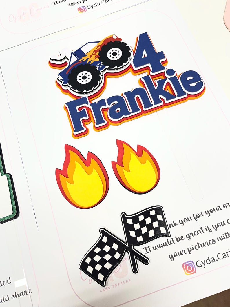 May include: A layered paper cake topper set featuring a blue monster truck with orange flames, the number 4, and the name Frankie in blue letters. The set includes two yellow and red flame cutouts and two crossed black and white checkered flags.