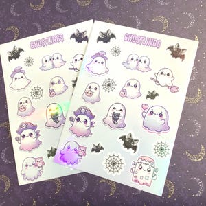 Puede incluir: Dos hojas de pegatinas holográficas con simpáticos personajes de fantasmas, murciélagos y telarañas. Las pegatinas tienen una combinación de colores pastel y el texto "Ghostlings" está impreso en la parte superior. El fondo es un patrón púrpura con lunas.