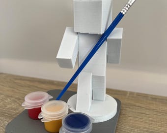 Könnte beinhalten: Eine weiße 3D-gedruckte Figur einer Person, die einen blauen Pinsel hält. Die Figur steht auf einem grauen Sockel mit drei kleinen Farbtöpfen: rot, gelb und blau.