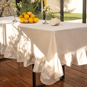May include: A rectangular cream-colored tablecloth with a ruffled edge, draped over a black metal table. The table is set with a plate of lemons and floral arrangements in silver vases, creating an elegant dining scene.