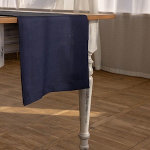 May include: A dark blue table runner on a white table with a wooden top. The table has a single turned leg visible in the image. The table runner is made of a soft, textured fabric.