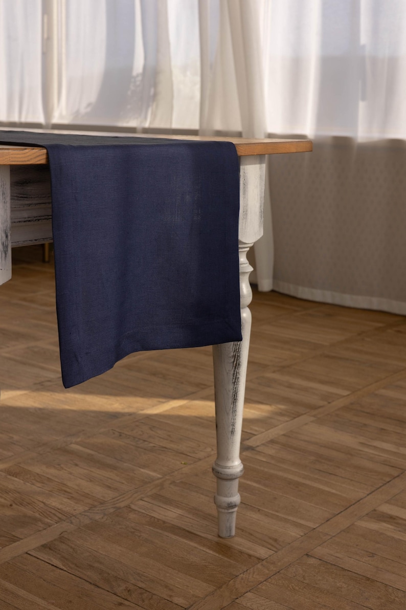 May include: A dark blue table runner on a white table with a wooden top. The table has a single turned leg visible in the image. The table runner is made of a soft, textured fabric.