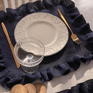 Puede incluir: Un mantel individual de lino azul marino con borde festoneado, un plato de cer&aacute;mica blanca con un dise&ntilde;o decorativo, una copa de vidrio transparente y cubiertos de oro. El mantel individual est&aacute; sobre un mantel de lino blanco. Hay tres posavasos de madera en la mesa.