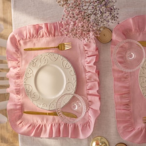 Puede incluir: Dos manteles individuales de lino rosa con volantes, con platos blancos y cubiertos dorados. Los manteles individuales est&aacute;n colocados sobre un mantel de lino beige claro.
