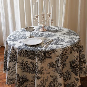 Puede incluir: Una mesa redonda cubierta con un mantel color crema con un patrón toile de jouy negro. Sobre la mesa hay un candelabro de plata de cinco brazos con velas blancas, un pequeño cuenco de plata con pie, un plato de cerámica blanca y dos tenedores.