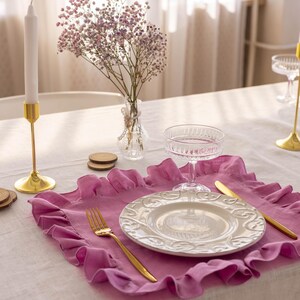 Puede incluir: Un mantel individual de lino rosa con volantes con un plato blanco, un tenedor y un cuchillo dorados, y una copa de vidrio transparente sobre un mantel blanco. Un peque&ntilde;o jarr&oacute;n con flores rosas est&aacute; en el fondo.