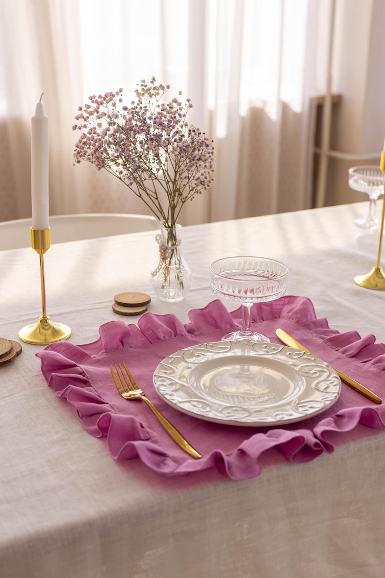Puede incluir: Un mantel individual de lino rosa con volantes con un plato blanco, un tenedor y un cuchillo dorados, y una copa de vidrio transparente sobre un mantel blanco. Un peque&ntilde;o jarr&oacute;n con flores rosas est&aacute; en el fondo.