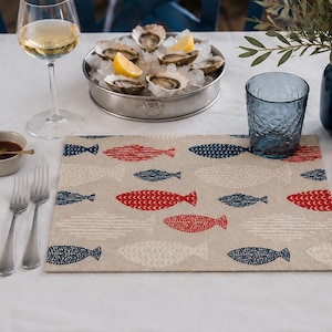 May include: A rectangular placemat featuring a repeating pattern of stylized fish in red, navy blue, and off-white on a light beige background. The fish are rendered with small dot and dash textures, creating a simple, repetitive design across the fabric.