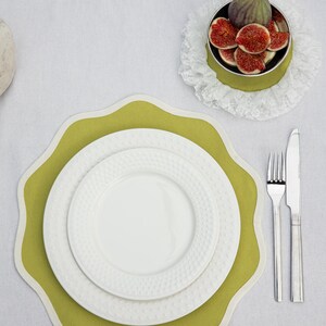 May include: A table setting featuring a green placemat with a scalloped edge, topped with white plates. A small bowl of figs sits on a lace doily. Silverware, including a fork and knife, is placed to the side.