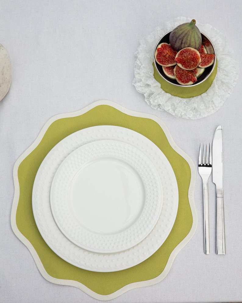 May include: A table setting featuring a green placemat with a scalloped edge, topped with white plates. A small bowl of figs sits on a lace doily. Silverware, including a fork and knife, is placed to the side.