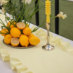 May include: A decorative table setting featuring a silver candlestick with a yellow candle, a silver platter of lemons, and a pale yellow table runner with a ruffled edge. White flowers and greenery add a fresh touch.