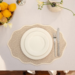 Op de afbeelding: Een tafelsetting met een beige bloemen placemat, witte borden en bestek. Een kom met citroenen en een kleine vaas met bloemen staan ook op de tafel, op een wit tafelkleed.