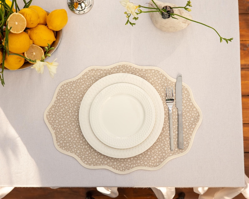 Op de afbeelding: Een tafelsetting met een beige bloemen placemat, witte borden en bestek. Een kom met citroenen en een kleine vaas met bloemen staan ook op de tafel, op een wit tafelkleed.
