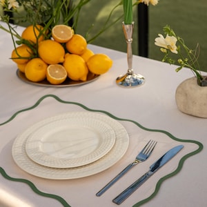 Pode incluir: Uma mesa posta com uma toalha branca, com um prato, um prato menor e talheres. Um sousplat com borda verde está sob os pratos. Um castiçal de prata com uma vela verde, uma tigela de limões e flores completam a cena.