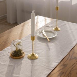 May include: Two gold candle holders with white candles on a wooden table with a white linen tablecloth. A small glass cloche with a plant inside sits on the table.