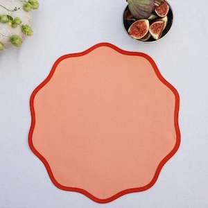 May include: A peach-colored placemat with a scalloped edge in a darker shade of red. The placemat is on a white surface, with a small bowl of figs and a decorative floral arrangement in the background.