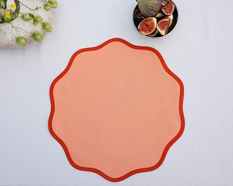 May include: A peach-colored placemat with a scalloped edge in a darker shade of red. The placemat is on a white surface, with a small bowl of figs and a decorative floral arrangement in the background.