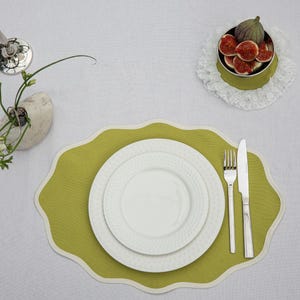 Op de afbeelding: Bovenaanzicht van een tafelsetting met een groene placemat, witte borden en bestek. Een kleine kom met vijgen staat op een kanten kleedje. Een kleine vaas met bloemen is ook aanwezig.